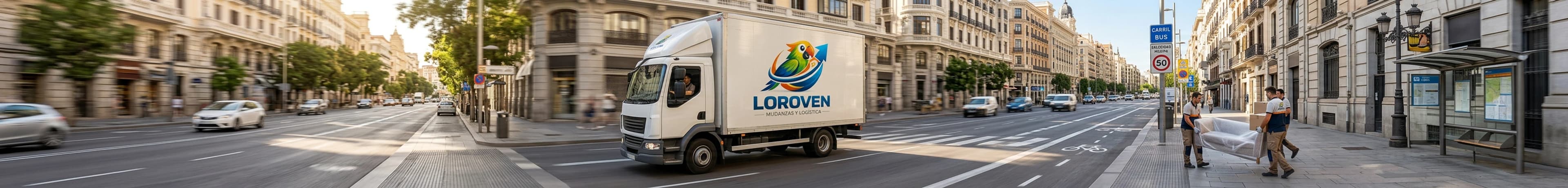Camión de mudanzas de Loroven circulando por una avenida mientras dos operarios trasladan un sofá embalado y cajas hasta el portal.